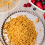 Crumbled graham cracker crust in a tart pan, with a bowl of berries and extra crust crumbs on a wooden surface.