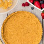 A tart pan filled with a pressed graham cracker crust sits on a wooden surface next to a bowl of berries and a partially empty mixing bowl with leftover crumbs.