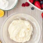 A glass bowl of whipped cream cheese mixture on a wooden surface, with a bowl of whipped cream, a cut lemon, and assorted fresh berries nearby.