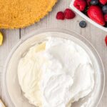 A bowl with whipped cream mixture, a graham cracker pie crust, and a bowl of mixed berries on a wooden surface.