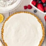 A cheesecake tart with a graham cracker crust sits on a wooden surface next to a bowl of mixed berries and an empty mixing bowl.