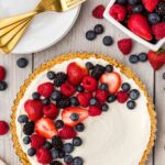 A cheesecake tart partially topped with fresh strawberries, raspberries, blueberries, and blackberries, next to plates, gold forks, and a bowl of assorted berries on a wooden table.