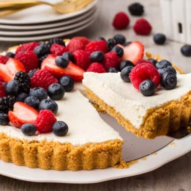 A tart with a crumb crust and creamy filling, topped with strawberries, raspberries, blueberries, and blackberries, with one slice being served on a spatula.