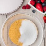 A glass bowl with graham cracker crumbs and sugar sits on a wooden surface next to fresh berries in a dish and a fluted tart pan.
