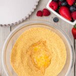 A glass bowl with graham cracker crumbs and melted butter, next to a tart pan, a dish of mixed berries, and a few scattered strawberries and raspberries on a wooden surface.