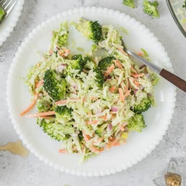 A plate of broccoli slaw with shredded carrots, broccoli florets, and creamy dressing, with a fork resting on the plate. A bowl of dressing and seasonings are nearby.