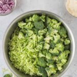A metal bowl filled with broccoli florets and shredded broccoli, surrounded by small bowls of shredded carrot, chopped red onion, dressing, and mayonnaise on a light surface.