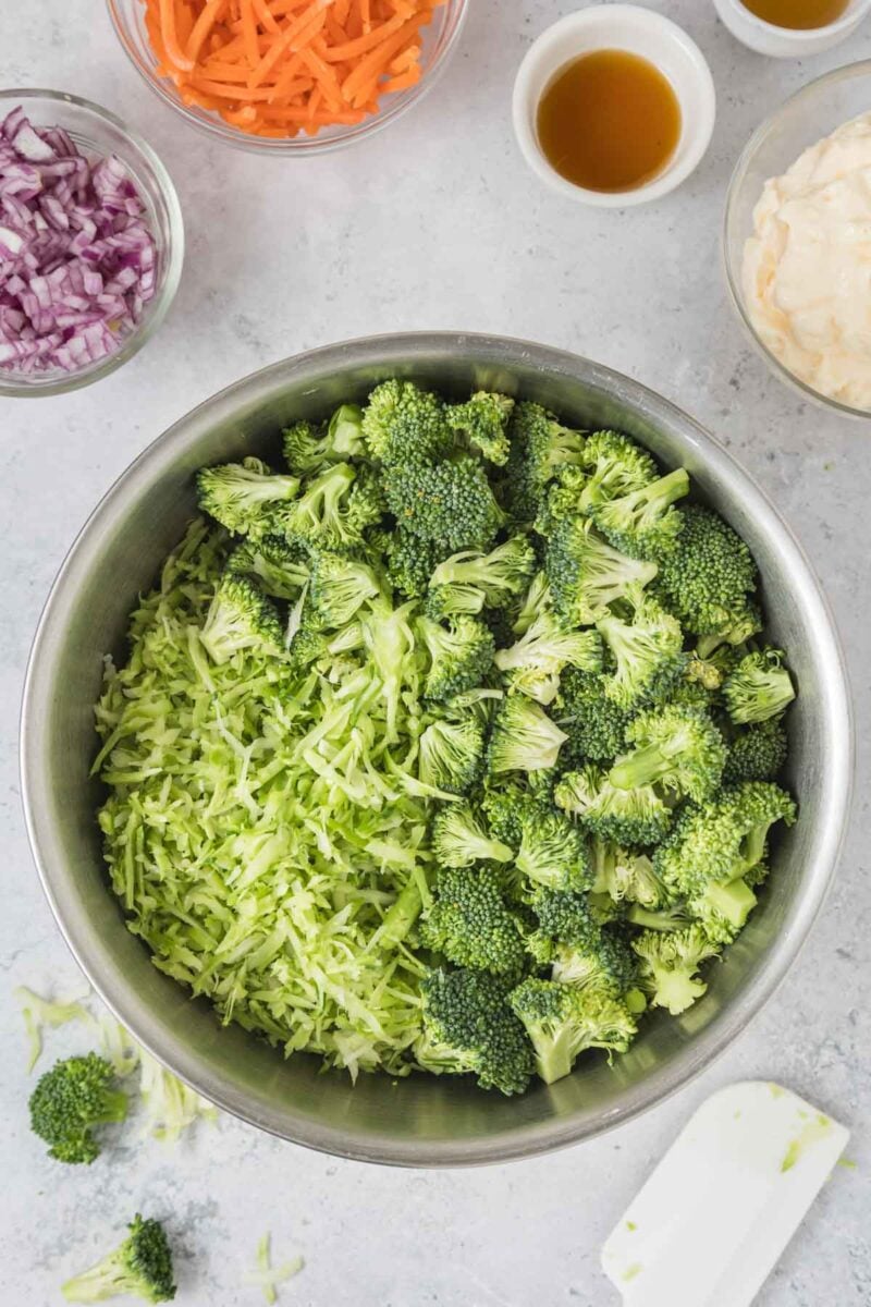 A metal bowl filled with broccoli florets and shredded broccoli, surrounded by small bowls of shredded carrot, chopped red onion, dressing, and mayonnaise on a light surface.