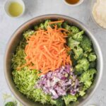 A metal bowl contains chopped broccoli, shredded carrots, diced red onions, and grated zucchini. Various small bowls with ingredients are arranged around it on a light surface.
