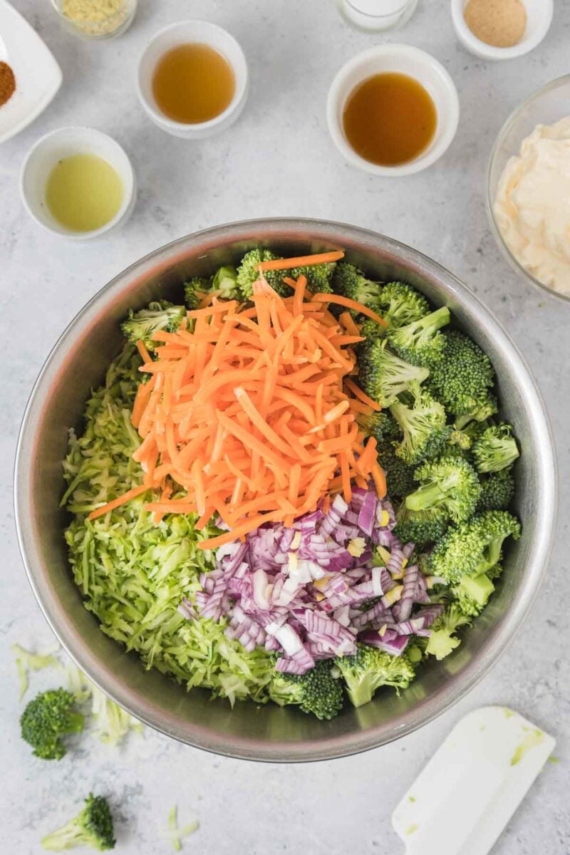 A metal bowl contains chopped broccoli, shredded carrots, diced red onions, and grated zucchini. Various small bowls with ingredients are arranged around it on a light surface.