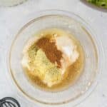 A glass bowl contains mayonnaise, spices, and vinegar mixture, ready to be whisked. Shredded cabbage and a lime are visible nearby on a light countertop.