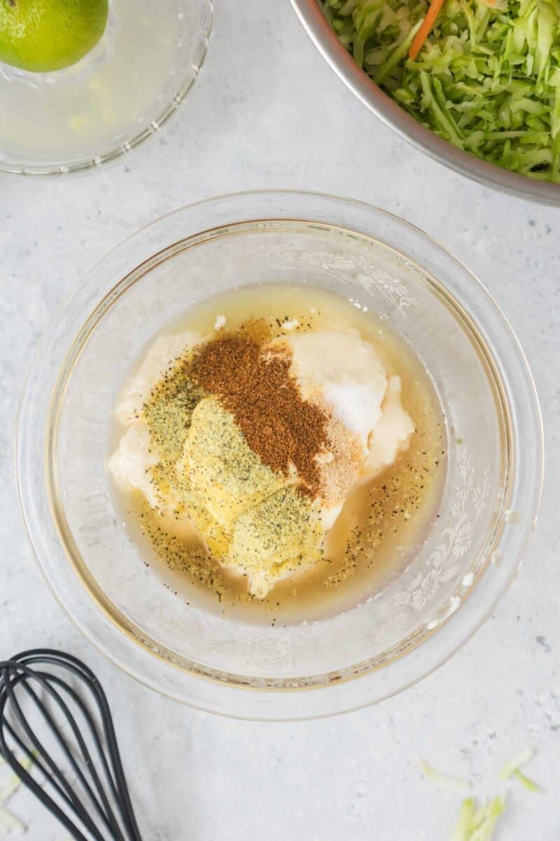 A glass bowl contains mayonnaise, spices, and vinegar mixture, ready to be whisked. Shredded cabbage and a lime are visible nearby on a light countertop.