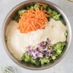A stainless steel bowl contains chopped broccoli, shredded carrots, diced red onion, and a creamy dressing, ready to be mixed. A whisk and lime half are nearby on the countertop.