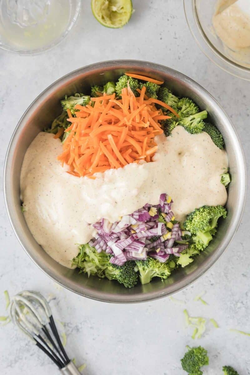 A stainless steel bowl contains chopped broccoli, shredded carrots, diced red onion, and a creamy dressing, ready to be mixed. A whisk and lime half are nearby on the countertop.