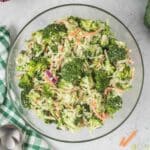 A glass bowl filled with broccoli slaw, containing broccoli florets, shredded carrots, and red onion, sits on a light surface beside a green-checked napkin and serving utensils.