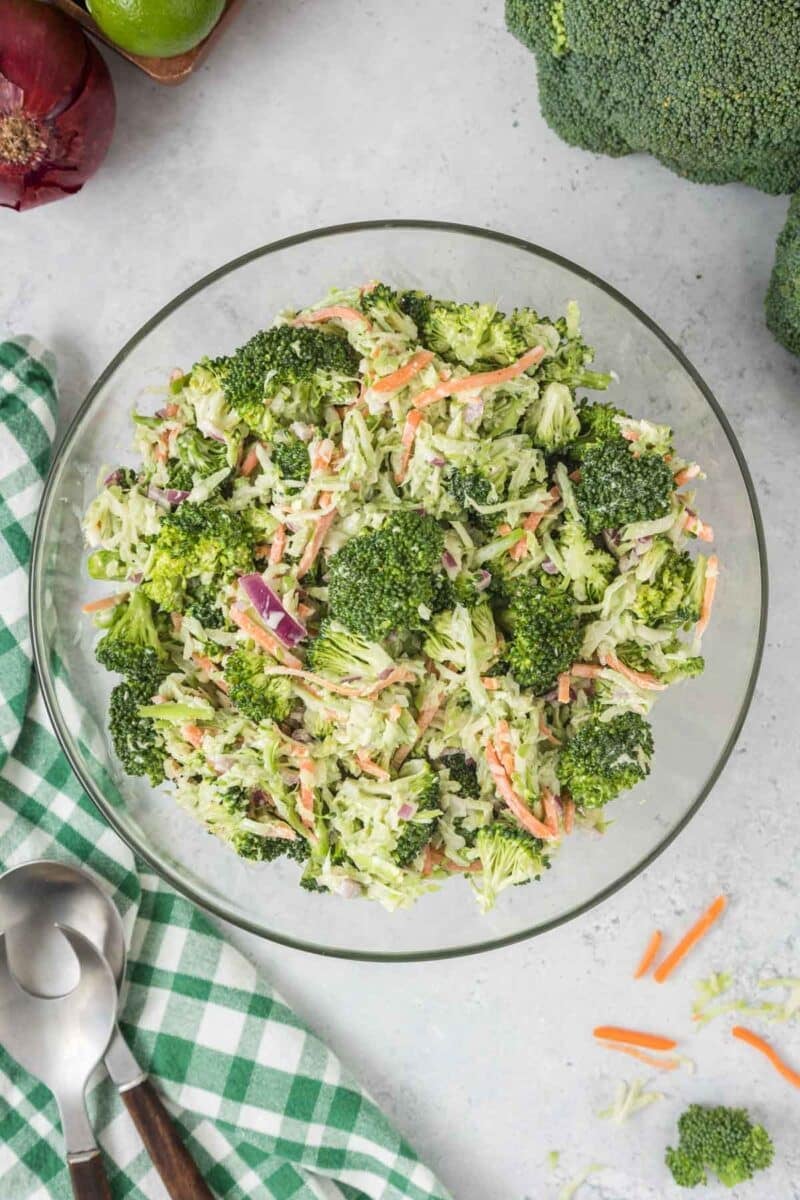 A glass bowl filled with broccoli slaw, containing broccoli florets, shredded carrots, and red onion, sits on a light surface beside a green-checked napkin and serving utensils.