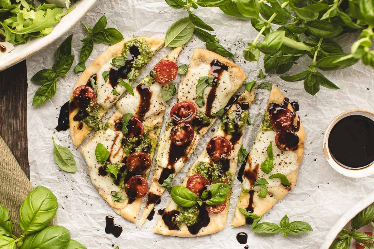 Caprese flatbread topped with melted cheese, cherry tomatoes, anchovies, basil leaves, and a drizzle of balsamic glaze, surrounded by fresh basil and a bowl of sauce.