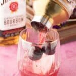 A hand pours a pink cocktail from a shaker into a glass with ice and cherries; a bottle of Jim Beam bourbon is in the background.