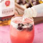 A clear liquid is being poured into a glass of red cocktail with ice and cherries. A Jim Beam bourbon bottle and cherries are visible in the background.