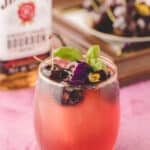 A glass of cherry bourbon smash cocktail garnished with cherries, edible flowers, and basil sits in front of a bottle of Jim Beam bourbon and a plate of cherries on a pink surface.