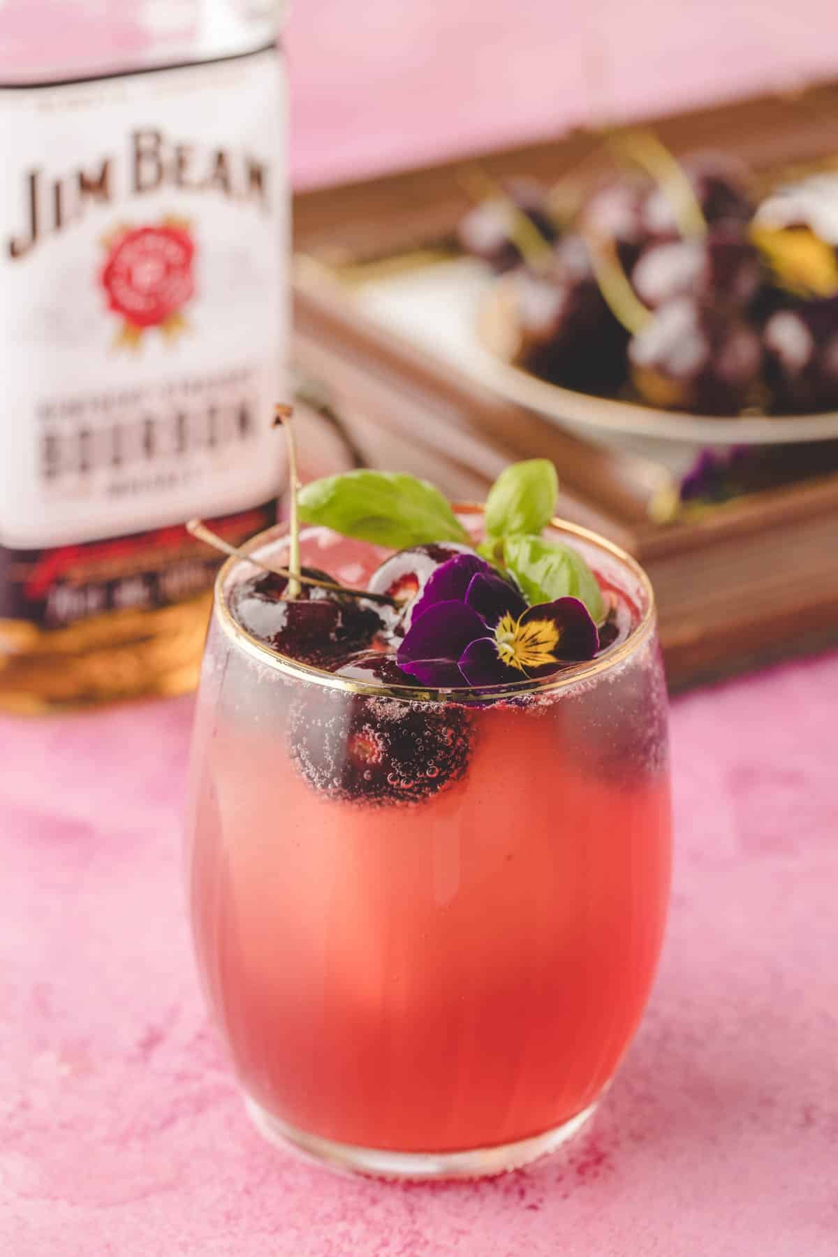 A glass of cherry bourbon smash cocktail garnished with cherries, edible flowers, and basil sits in front of a bottle of Jim Beam bourbon and a plate of cherries on a pink surface.