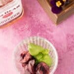 A glass with sliced dark cherries and fresh basil leaves on a pink surface, next to a bottle of Jim Beam bourbon and a wooden tray with a purple flower.