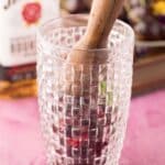 A wooden muddler pressing berries in a textured glass, with a bottle of Jim Beam bourbon visible in the background.