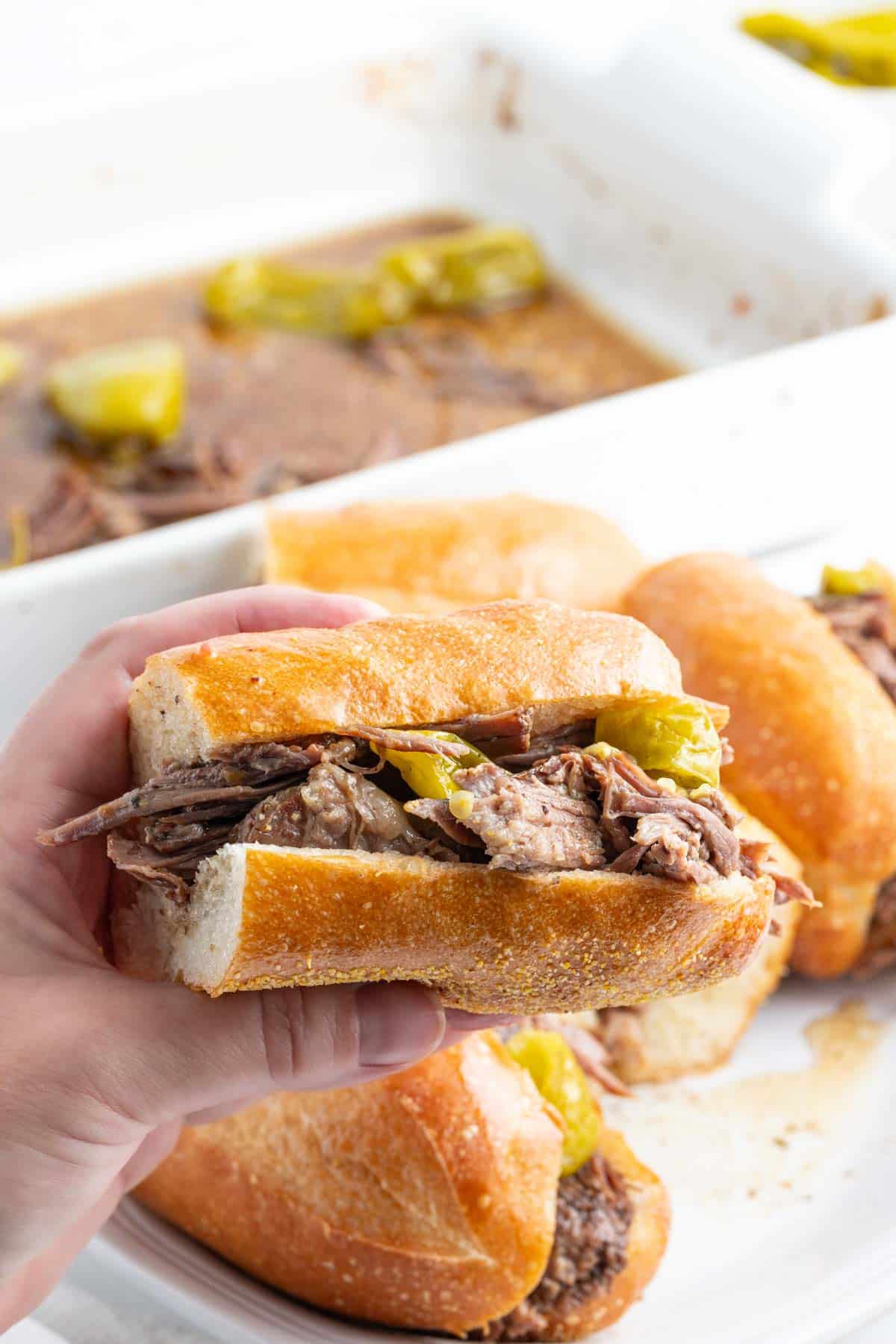 A hand holding a sandwich filled with chicago italian beef and pepperoncini, with more sandwiches and a baking dish in the background.