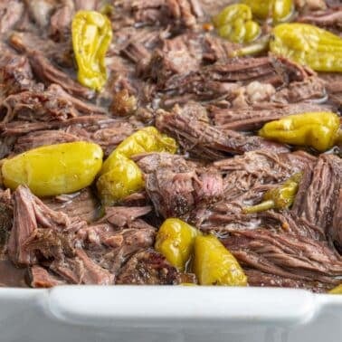 Shredded pot roast with yellow pepperoncini peppers in a white baking dish.