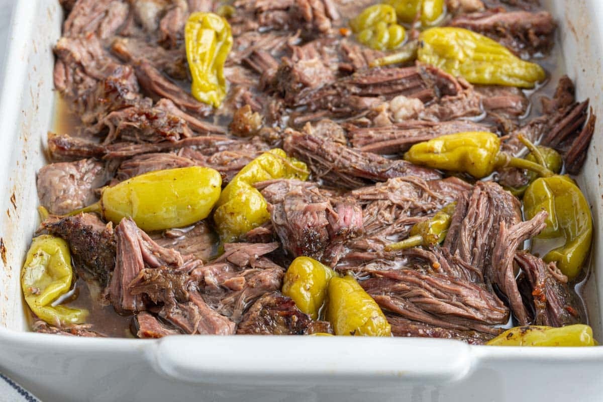 Shredded pot roast with yellow pepperoncini peppers in a white baking dish.