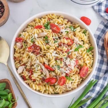 A bowl of rotini pasta salad with cherry tomatoes, shredded cheese, herbs, and chicken sits on a table with a gold spoon, fresh basil, green onions, and a bowl of grape tomatoes nearby.