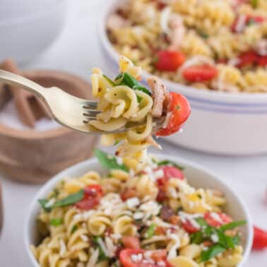 A fork holds a serving of chicken and bacon pasta salad with rotini, cherry tomatoes, spinach, and shredded cheese above a bowl of the same dish.