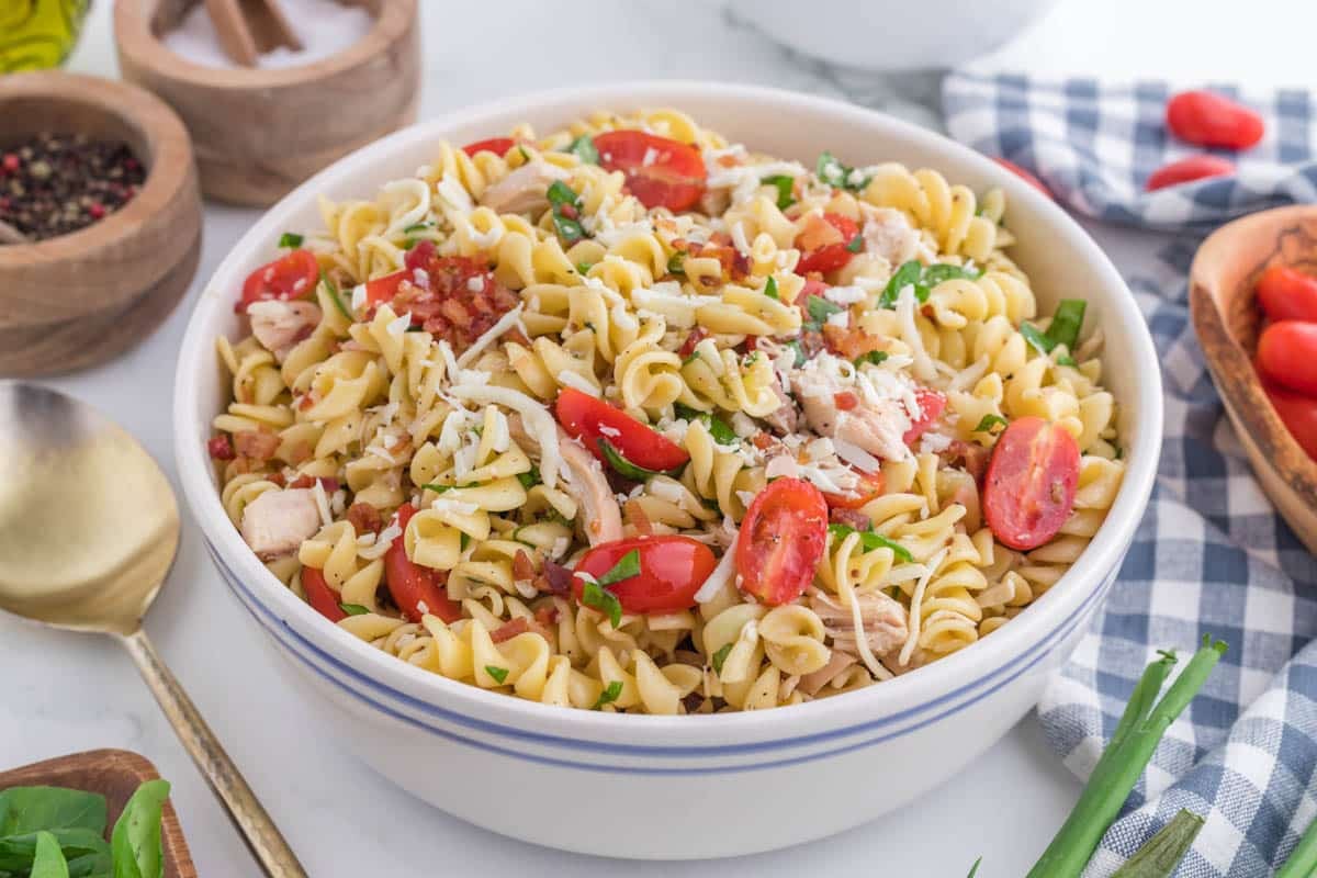 A bowl of chicken bacon pasta salad with cherry tomatoes, shredded chicken, parsley, and grated cheese, placed on a white surface with a gold spoon and seasonings nearby.