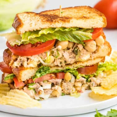 A chickpea tuna salad sandwich with lettuce and tomato, cut in half and stacked, served on a plate with potato chips.