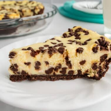 A slice of chocolate chip cheesecake on a white plate, with a glass of milk and the rest of the cheesecake in the background.