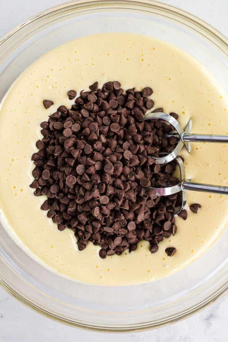 A hand mixer and chocolate chips in a bowl of light yellow batter on a marble surface, with an empty glass bowl and a chocolate pie crust nearby.
