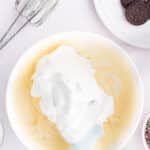 A bowl with whipped cream being mixed into a yellow batter, surrounded by a hand mixer, cookies, mini chocolate chips, and small bowls of ingredients.