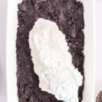 A baking dish with a layer of crushed chocolate cookies being topped with a layer of white cream mixture using a spatula.