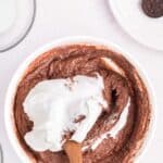 A bowl of chocolate batter being mixed with whipped cream using a wooden spoon, surrounded by small bowls of ingredients and chocolate cookies.