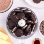 A food processor filled with chocolate sandwich cookies sits on a counter surrounded by butter, cocoa powder, chocolate chips, vanilla extract, and extra cookies on a plate.