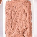 A rectangular white baking dish filled with chocolate batter being spread with a spoon, surrounded by bowls of ingredients.