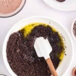 A bowl of crushed chocolate cookies mixed with melted butter, a spatula, and surrounding ingredients including milk, cocoa powder, whole cookies, and chocolate chips.