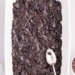 A rectangular baking dish with a layer of crushed chocolate cookies being spread with a spoon; ingredients surround the dish.