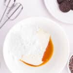 A white bowl contains cream cheese, powdered sugar, and vanilla extract. Surrounding the bowl are beaters, chocolate sandwich cookies, and small bowls with other ingredients.