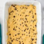 A baking dish filled with unbaked chocolate chip cookie dough, spread evenly on parchment paper, sits on a marble countertop next to a plate of sandwich cookies.