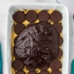 A baking dish lined with Oreos and cookie dough, topped with a layer of chocolate brownie batter, sits on a white surface.