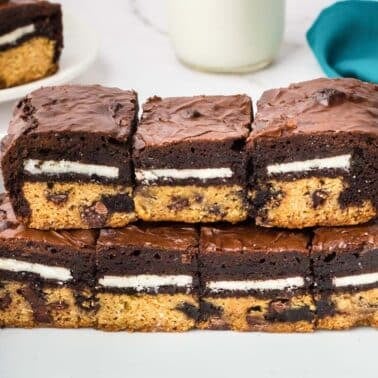 Rectangular Easy slutty brownies with layers of chocolate chip cookie, Oreo cookie, and brownie, arranged on a white tray with a teal napkin and milk in the background.