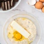 A bowl with flour, chocolate chips, a stick of butter, and an egg on a countertop next to a bowl of cookies and a dish of eggs.