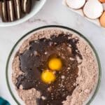 A glass bowl with chocolate cake mix, two cracked eggs, and oil, next to a plate of sandwich cookies and a dish with whole and cracked eggs on a marble surface.