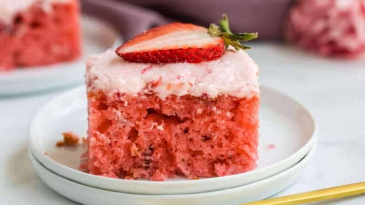 A slice of pink strawberry cake with frosting and a sliced strawberry on top, served on a white plate—perfect for anyone who loves flavorful desserts.